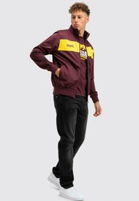 Bordó zipzáras dzseki sárga vízszintes csíkokkal, bordás mandzsettákkal, két oldalsó zsebbel, fekete póló fölött viselve, fekete farmerral és fehér sportcipővel párosítva.