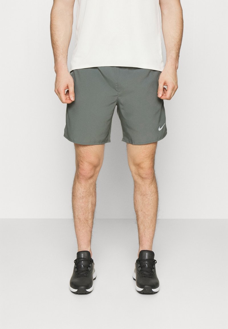 Homme portant un short de sport Nike gris, des chaussures de course noires et une chemise blanche, debout devant un fond uni.