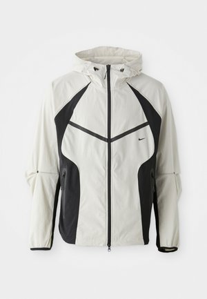 Veste à capuche légère en crème avec des panneaux latéraux noirs, fermeture éclair et poignets élastiques. Dotée d'une petite poche zippée et d'accents réfléchissants.