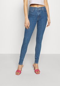 Vaqueros ajustados en denim azul medio, con un diseño clásico de cinco bolsillos, detalles de costura y una textura suave. Combinados con sandalias deslizantes rosas.