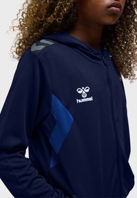 Mørkeblå hættetrøje lavet af tekstureret stof med lynlåslukning, blå accenter på siden og et logo på brystet. Hættedesign.