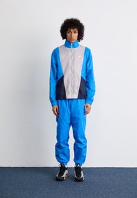 Un survêtement comprenant une veste bleue avec des panneaux gris et marine, un pantalon bleu assorti, et des baskets noires avec des accents. Texture lisse et sportive.