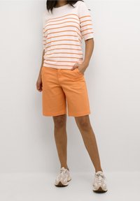 Vrouw in een wit en oranje gestreepte korte mouwen top, oranje knielange shorts en witte veterschoenen staat tegen een witte achtergrond.
