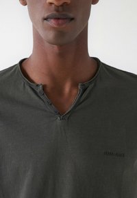 Dunkelgrünes Kurzarmhemd mit V-Ausschnitt und drei Knöpfen am Ausschnitt. Mit "Salsa Jeans" in den Stoff eingeprägt. Glatte Textur.