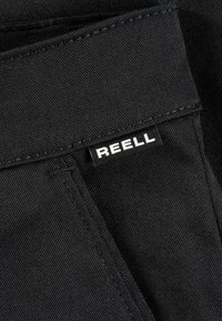 Black denim fabric with a visible texture; features a rectangular label with the brand name "REELL" in white text on a black background.