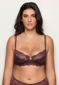 Donna dai capelli scuri che indossa un set coordinato di reggiseno e slip in pizzo viola scuro, in piedi contro uno sfondo chiaro e semplice.