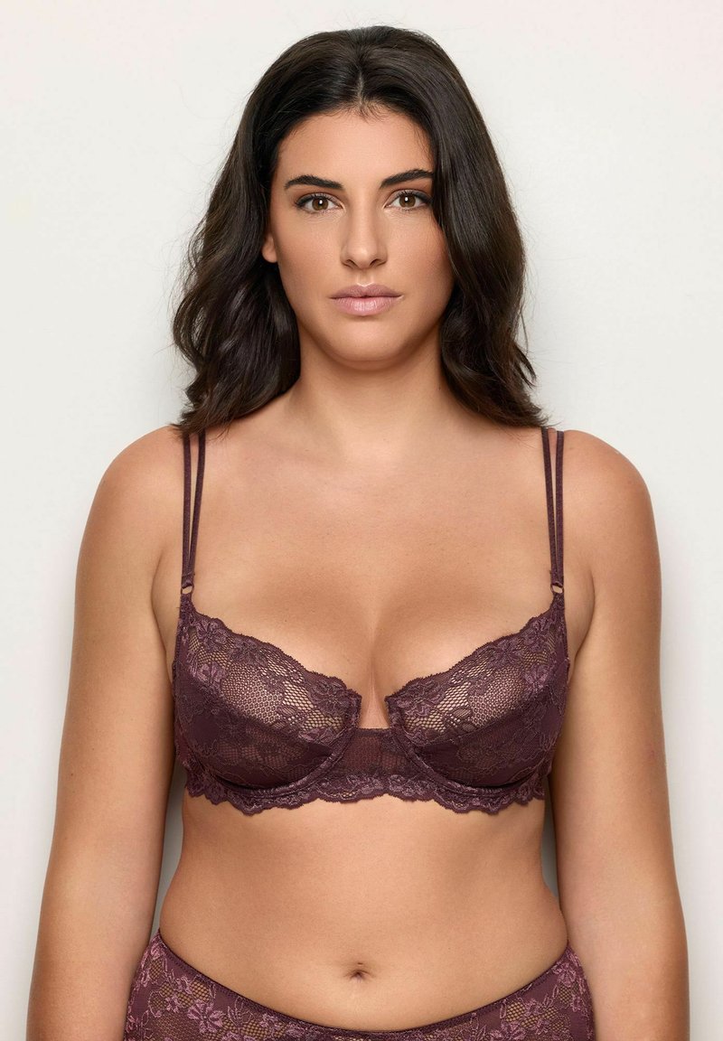 Donna dai capelli scuri che indossa un set coordinato di reggiseno e slip in pizzo viola scuro, in piedi contro uno sfondo chiaro e semplice.
