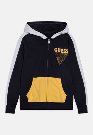 Granatowa bluza z kapturem na suwak, z białą podszewką kaptura, białymi paskami na rękawach, żółtymi kieszeniami z przodu oraz żółtym logo „GUESS” na piersi.