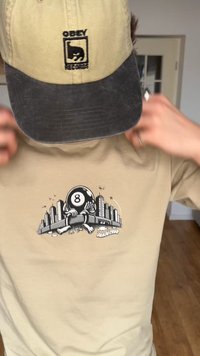 Osoba nosiaca béžovú a čiernu čiapku s logom "Obey Records" a béžové tričko s postavičkou osemky, ktorá drží panorámu mesta.