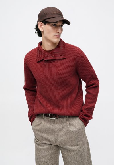 Red knitted sweater with a wide collar and buttons, paired with textured beige trousers and a dark brown cap.