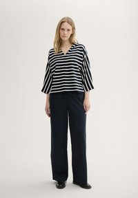 Gestreiftes, marineblaues und weißes V-Ausschnitt-Oberteil mit weiten Ärmeln, kombiniert mit hochtaillierten, dunkelmarineblauen Hosen und schwarzen Schuhen. Glatte Stofftextur.