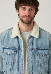 Lichtblauwe denimjack met een witte sherpa-kraag, drukknoopsluiting, twee borstzakken en een rode Levi's-tag op de linkse zak.
