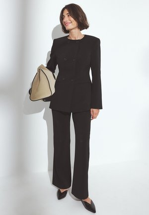 Woman with short brown hair wears tailored black suit and pointed black shoes, holding large beige handbag against a white background.
