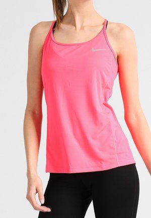 Frau trägt ein leuchtend pinkes ärmelloses Nike-Sporttop und schwarze Leggings, steht vor einem einfarbigen Hintergrund.