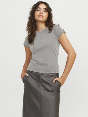 Femme aux cheveux bouclés portant un t-shirt gris moulant et une jupe en cuir foncé, debout les mains dans les poches devant un fond uni.
