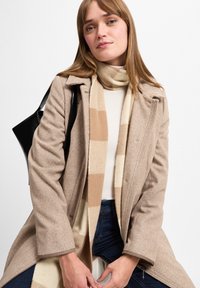 Beige uldfrakke med en hørmønster, parret med et stribet beige og cremefarvet tørklæde, suppleret med mørke jeans og en sort taske.