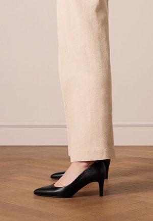Black leather high-heeled shoes with a pointed toe design, paired with light beige wide-leg pants, on a wooden floor.