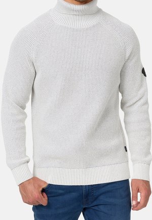 Man wearing white textured knit turtleneck sweater and blue jeans with one hand in pocket.