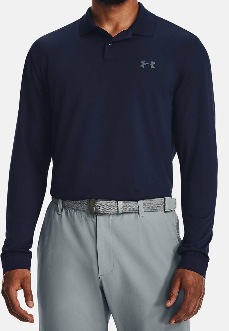 Homme portant un polo à manches longues bleu marine avec un petit logo et un pantalon gris clair avec une ceinture tissée.