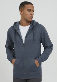 Sweat à capuche zippé en tissu gris foncé avec poches avant, poignets et taille côtelés. Texture lisse avec cordons de serrage sur la capuche.