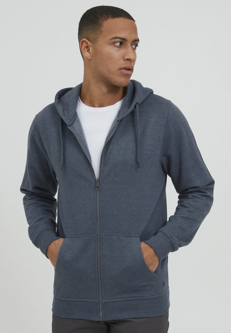 Sweat à capuche zippé en tissu gris foncé avec poches avant, poignets et taille côtelés. Texture lisse avec cordons de serrage sur la capuche.