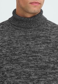 Homme portant un pull en maille chinée noir et gris foncé, montrant le cou, le menton et la partie supérieure de la poitrine.