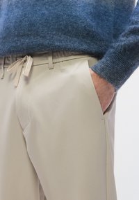 Beiget housut, joissa on sileä pinta, joustava vyötärönauha ja kiristysnauha. Yhdistetty siniseen, pehmeästä villasta valmistettuun neulepaitaan.