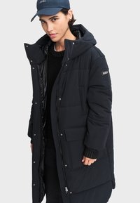 Padded čierna parka s vysokým golierom, zipsom, bočnými vreckami a odnímateľnou kapucňou. Vlastnosti zahŕňajú hladký vonkajší povrch a izolovanú podšívku.