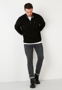 Pullover in pile nero con zip a quarto, maniche lunghe e un piccolo logo sul petto. Indossato con jeans aderenti grigi e stivaletti neri.