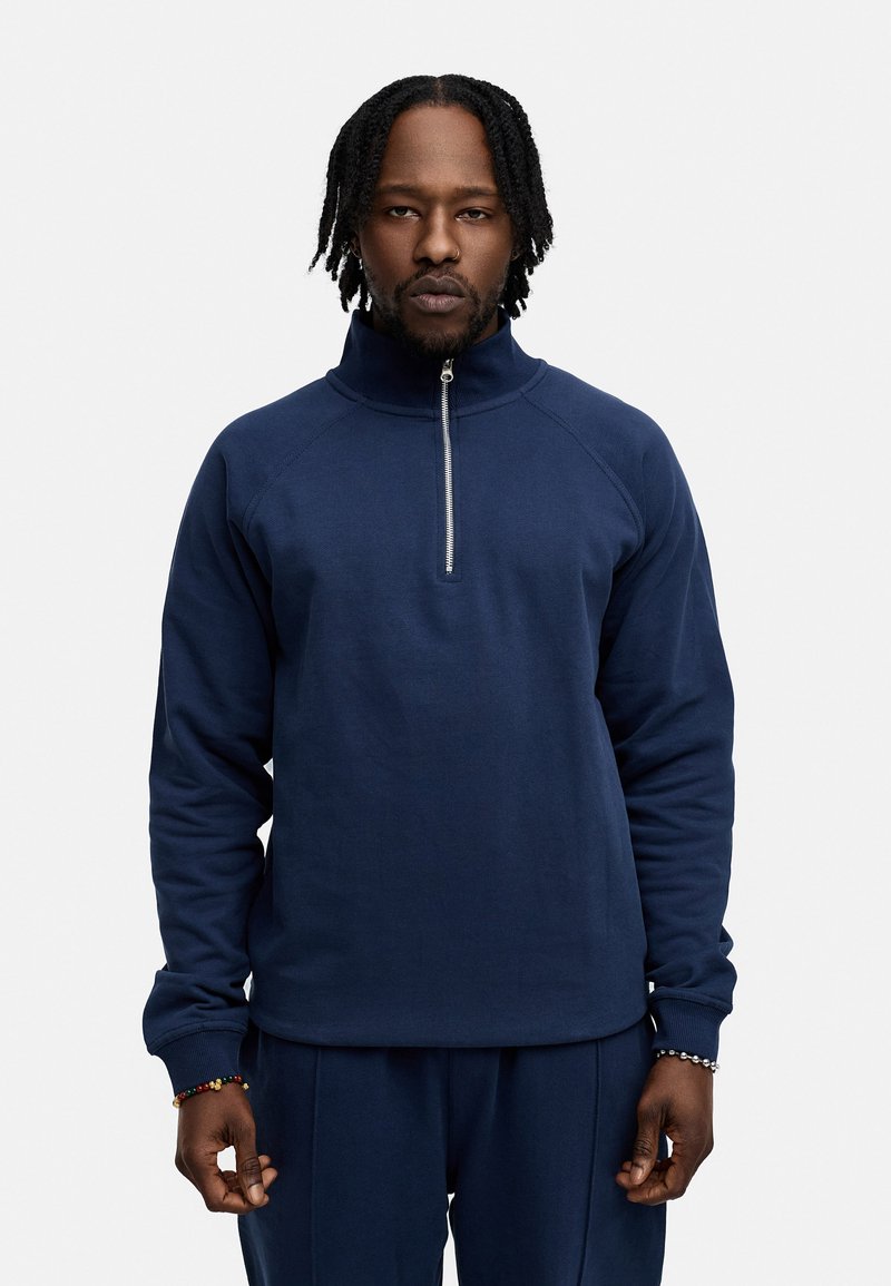 Navy zip-up pullover made of textured fabric, featuring a high collar and raglan sleeves; worn with matching joggers.