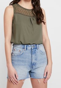 Top sans manches vert olive avec un empiècement transparent à pois et des détails plissés, associé à un short en denim bleu clair taille haute avec des bords effilochés.