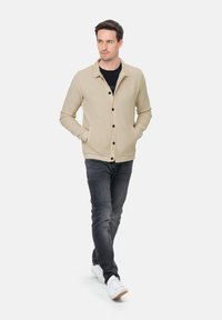 Beige Blazer mit strukturiertem Muster, getragen über einem schwarzen T-Shirt, kombiniert mit dunklen Jeans und weißen Sneakers.