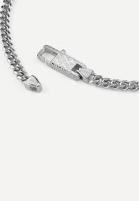 Bracelet en chaîne en argent avec un fermoir texturé, présentant une étiquette triangulaire avec le logo "GUESS". Maillons lisses entrelacés, design moderne.
