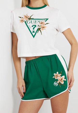 Vrouw draagt een wit cropped T-shirt met groen Guess-logo en bloemrijke borduursels, gecombineerd met groene sportshorts met een bijpassend bloemmotief.