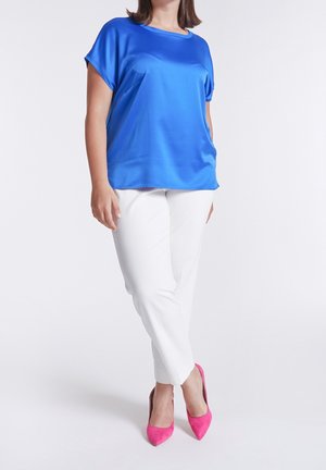 Mujer con blusa de manga corta color azul brillante, pantalones blancos ajustados y tacones altos puntiagudos rosas, de pie con las piernas cruzadas.