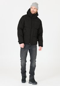 Svart puffjacka med hög krage, frontdragkedja och två sidofickor. Bärs med grå jeans och svarta vandringskängor, mot en ljus bakgrund.