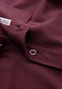 Primer plano del cuello de una camisa de tela texturizada color granate con un botón abrochado y una etiqueta blanca parcialmente visible.
