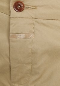 Beżowa bawełniana tkanina o gładkiej fakturze. Zawiera brązowy guzik i małą metkę z napisem "CLOSED" po boku. Widoczne detale szwów.