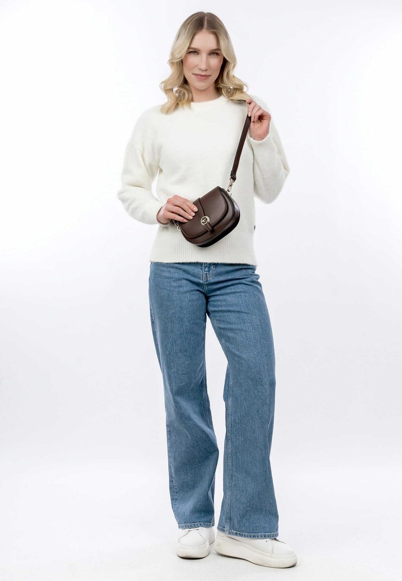 Borsa a spalla marrone con forma arrotondata, in pelle, con accessori in argento e chiusura a pattina. Modella che indossa un maglione bianco e jeans blu.