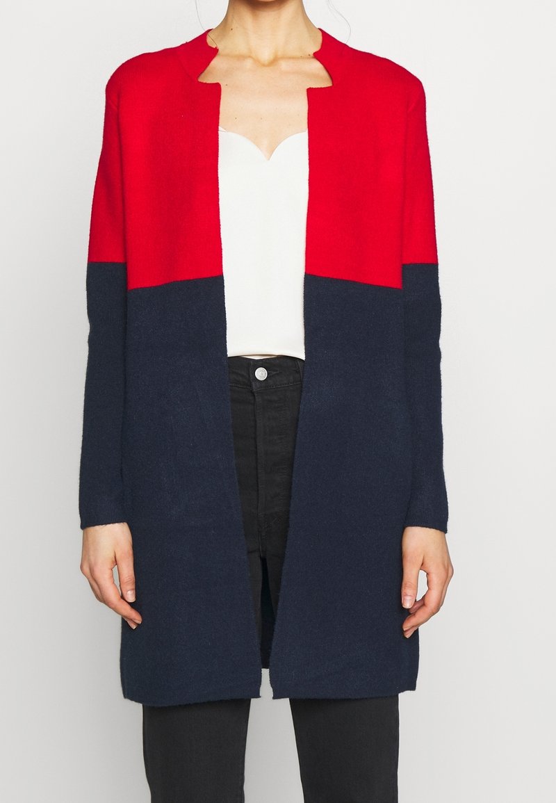 Long cardigan à blocs de couleur rouge et marine, devant ouvert, poignets côtelés et texture douce. Porté sur un haut blanc et un pantalon noir.