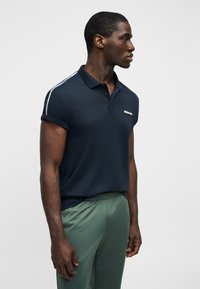 Άνδρας που φοράει μπλε σκούρο πόλο πουκάμισο με ανασηκωμένα μανίκια και πράσινο παντελόνι, στέκεται προφίλ μπροστά σε απλό φωτεινό φόντο.