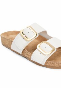 White sandal with two wide straps, each featuring a large gold buckle, and a cork footbed with a rubber sole.