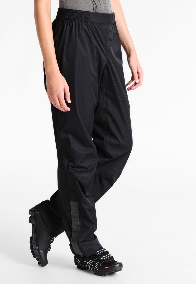 Personne portant un pantalon imperméable noir et des chaussures de cyclisme noires, debout avec un pied croisé devant l'autre.