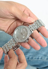 Srebrny zegarek na nadgarstek z teksturowanym cyferblatem, wskaźnikiem dnia i błyszczącymi akcentami. Bransoleta ze stali nierdzewnej o konstrukcji ogniw, trzymana w dłoni.