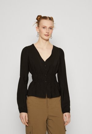 Black blouse with gathered waist and puffed long sleeves, featuring a deep V-neck and button closure. Worn with beige pants.