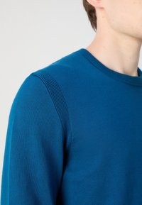 Gros plan d'une personne portant un pull en tricot bleu avec des détails côtelés sur l'épaule et un col rond.