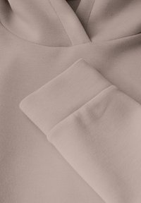 Tessuto del felpa rosa chiaro che mostra una morbida texture con un collo leggermente strutturato e polsini arrotolati. Dettagli di cucitura minimalista sono visibili.