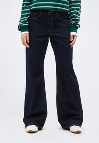 Άτομο που φοράει σκούρα μπλε τζιν παντελόνια με φαρδιά ποδαρίλα, λευκά αθλητικά παπούτσια και μπλούζα με μακριά μανίκια σε πράσινες και λευκές ρίγες, στέκεται μπροστά σε λευκό φόντο.