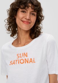 Biała koszulka z krótkim rękawem z pomarańczowym, teksturowanym napisem "SUN SATIONAL" na klatce piersiowej. Wykonana z miękkiego bawełnianego materiału, luźny krój.