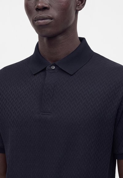 Gros plan d'une personne portant un polo texturé sombre avec un col et un bouton fermé.